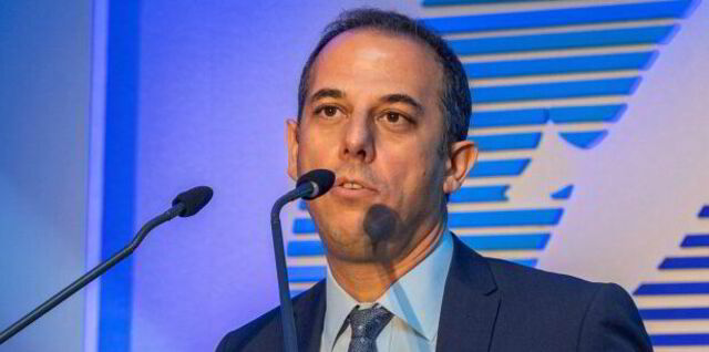 A man in a suit speaks into two microphones at a podium during an event, with a blue background behind him